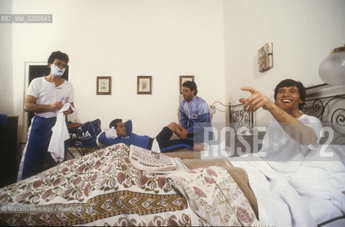 Italian National Singers Football Team, 1987. Singers Riccardo Fogli, Gianni Bella, masseur Nicola Mancuso, singer Gianni Morandi in a hotel room  during a tour of the talian National Singers Football Team / Nazionale italiana cantanti 1987. I cantanti Riccardo Fogli, Gianni Bella, il massaggiatore Nicola mancuso e Gianni Morandi in una stanza dalbergo durante una tournèe della Nazionale italiana cantanti - ©Marcello Mencarini/Rosebud2