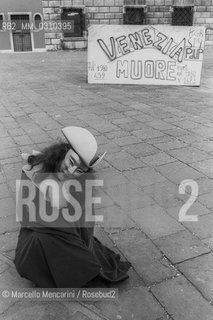 Venice Carnival 1982. Mask in front of a sign denouncing the negative trend of the population in the city because deaths rated exceed births / Carnevale di Venezia 1982. Una maschera davanti a un cartello che denuncia lo spopolamento della città, per il maggior numero di morti rispetto alle nascite - ©Marcello Mencarini/Rosebud2