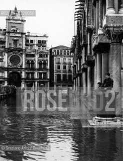 Venice,1957. Piazza San Marco. Ragazzo pesca appoggiato alla Basilica di San Marco durante lalta marea. Sullo sfondo le passerelle di emergenza. Young with a fishing pole in Venicès famed San Marco Square, inundated by exceptonally high tides. In background, pedestrians make their way over the flood waters on emergency catwalks. Acqua alta;alta marea; pesca; passerelle - La cessione dei diritti si intende per quanto di nostra competenza; non comprende invece le eventuali spese relative a diritti che potranno essere richiesti dagli Enti cui appartengono le opere riprese. Tali costi, ove neccessari, e l’espletamento di qualsivoglia pratica di richiesta permessi sono esclusivamente  a  carico del committente. © ARCHIVIO Graziano Arici/Rosebud2 